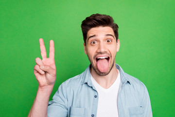 Closeup photo of amazing guy showing v-sign symbol sticking tongue out mouth wear casual denim shirt isolated over green color background