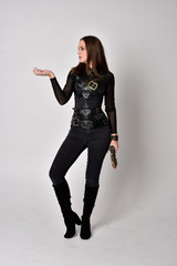 full length portrait of a pretty brunette woman wearing black leather fantasy costume with a gun. standing pose on a studio background.