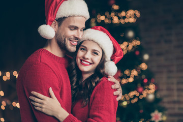 Closeup photo of overjoyed couple spending christmas time eve in decorated garland lights room near x-mas tree indoors wearing red pullovers and santa hats