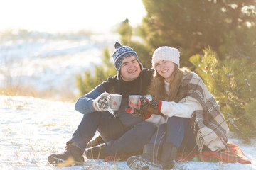 Fototapeta na wymiar Young couple in love drink a hot drink with marshmallows, sitting in the winter in the forest, tucked in warm, comfortable rugs and enjoy nature. They talk and laugh for a cup of coffee.