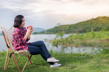 Naklejka na ściany i meble smart asian female travel in forest, she holding red ceramic cup and drinking hot coffee at lakeside , she sitting on seat, adventure activity and travel