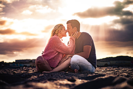 Romantic scenic moment and love concept with couple of young beautiful people hugging and kissing sit down on the ground with golden amazing sunset in backgorund