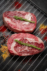 Two juicy raw rib eye steaks prepared for grilling with spices and herbs.