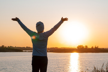 Fototapeta premium A young man, a hipster, stands on a lake at sunset, enjoys the view while traveling, desolation, enjoyment, harmony.