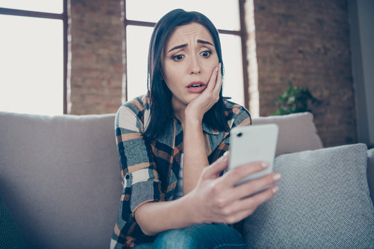 Photo of charming lady holding telephone hand not believe eyes reading new blog post negative comments sitting sofa wearing casual clothes apartment indoors