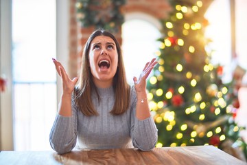 Young beautiful woman sitting at the table at home around christmas tree and decoration crazy and...