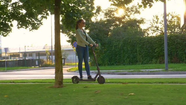 SLOW MOTION, LENS FLARE: Young Caucasian woman rides an e-scooter along a sidewalk in the modern city. Millennial girl riding a cool high tech electrical scooter through the city on a sunny evening.