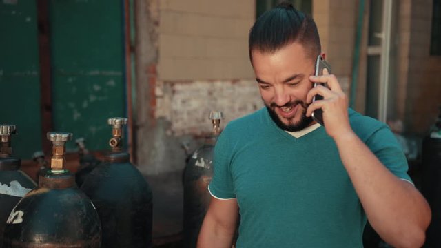 Young man with dark hair and beard, blue t-shirt holding smartphone, standing near old building, factory, talking on phone, showing thumbs up sign OK, considers gas cylinders