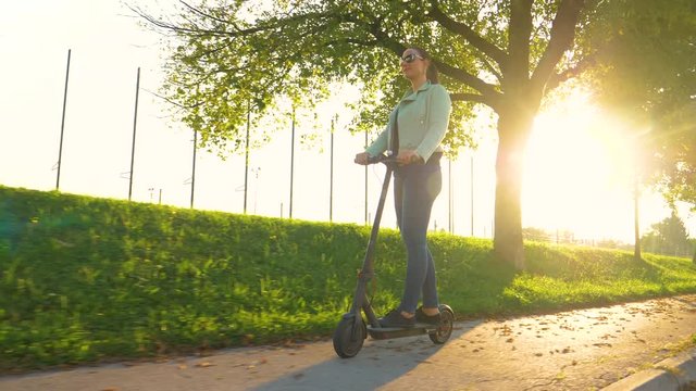 LOW ANGLE, SUN FLARE: Young woman in jeans rides a high tech e-scooter along an avenue in the autumn. Caucasian girl having fun riding an electric scooter down empty sidewalk on a sunny fall evening.
