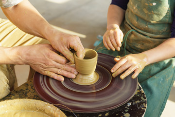 Fototapeta na wymiar The hands of an adult man show the child how to make a jug from a piece of clay on a Potters wheel