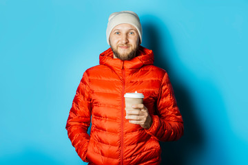 Man in a hat and a red jacket holds a cup of hot coffee on a blue background. Warming drinks,...