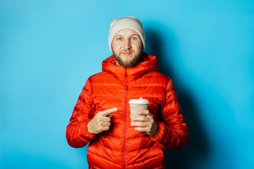 Man in a hat and a red jacket holds a cup of hot coffee on a blue background. Warming drinks,...