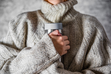 woman in a sweater holds a thermomug with a hot drink in her hands on gray wall background