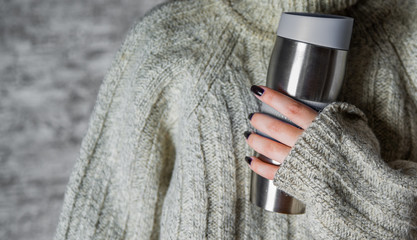 Fototapeta premium woman in a sweater holds a thermomug with a hot drink in her hands on gray wall background