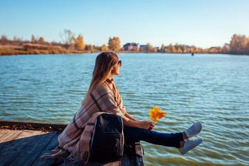 Traveler with backpack relaxing by autumn river at sunset. Young woman sitting on pier admiring...