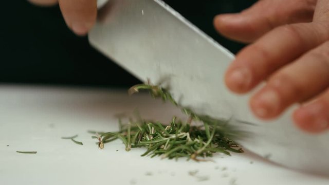 Close up of a professional chef in a Mediterranean restaurant chopping fresh rosemary on a board with a kitchen knife