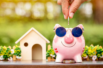 Businessman hand hold a coin is save to the piggy bank with growing a interest and wooden home model for family in the public park, Business investment and loan for real estate in the future concept.