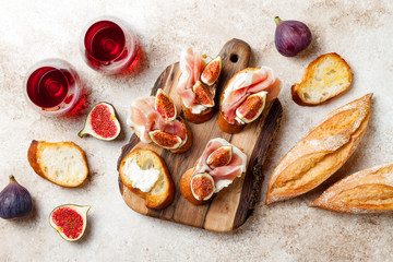 Crostini with prosciutto, cream cheese and figs on wooden board. Appetizers, antipasti snacks and red wine in glasses. Authentic traditional spanish tapas set. Light beige background. Top view