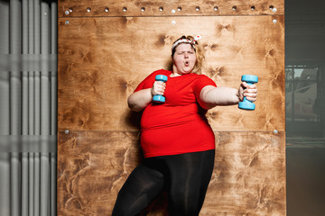 Funny fat girl dressed in the sportswear and with a bandage on her head stands with dumbbells on...