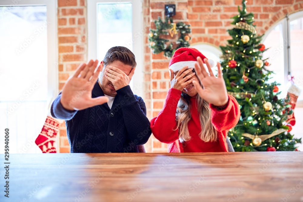 Sticker young couple wearing santa claus hat sitting on chair and table around christmas tree at home coveri