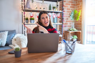 Beautiful woman sitting at the table working with laptop at home around christmas lights smiling and looking at the camera pointing with two hands and fingers to the side.