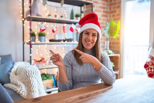 Young beautiful woman wearing christmas hat sitting at the table at home smiling and looking at the camera pointing with two hands and fingers to the side.