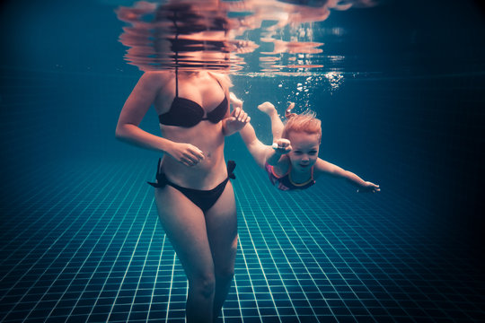 Happy child learning swimming underwater in swimming pool during diving training course with helping mother
