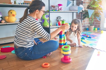 Beautiful teacher woman and toddler playing with building blocks around lots of toys at kindergarten