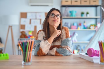 Young beautiful teacher woman wearing sweater and glasses sitting on desk at kindergarten Thinking concentrated about doubt with finger on chin and looking up wondering