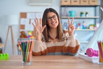 Young beautiful teacher woman wearing sweater and glasses sitting on desk at kindergarten showing...