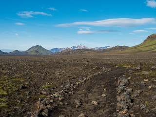 Icelandic lava desert landscape with footpath of Laugavegur hiking trail with view on Tindfjallajokull glacier mountains and green hills. Fjallabak Nature Reserve, Iceland. Summer blue sky