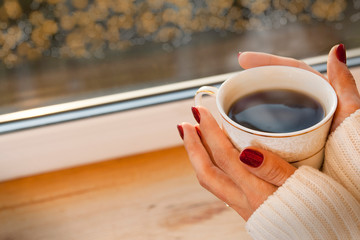 A girl in a sweater is standing by the window with a cup of coffee. Outside the window glare, bokeh