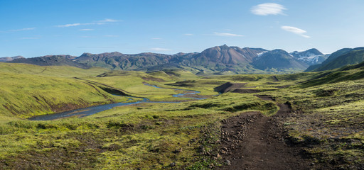 Panoramic landscape with blue river stream, green hills, snow-capped mountains, meadow and lush...