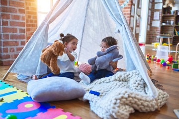 Adorable toddlers lying down over blanket inside tipi smiling and playing with doll at kindergarten