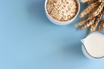 Oat milk in a jug and oats in a white ceramic bowl on a blue paper background. Alternative type of milk. Vegan substitute dairy milk.