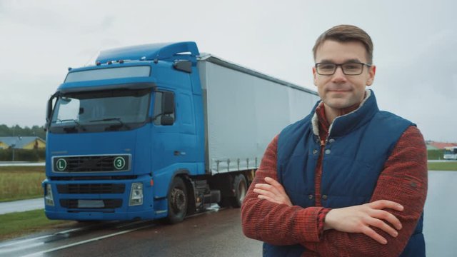 Professional Young Truck Driver Crosses Arms and Smiles. Behind Him Parked Blue Long Haul Semi-Truck with Cargo Trailer