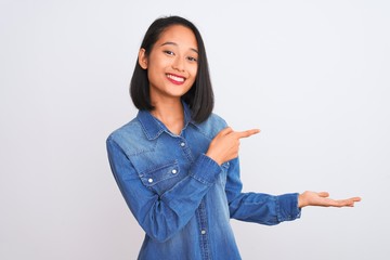 Young beautiful chinese woman wearing denim shirt standing over isolated white background amazed and smiling to the camera while presenting with hand and pointing with finger.