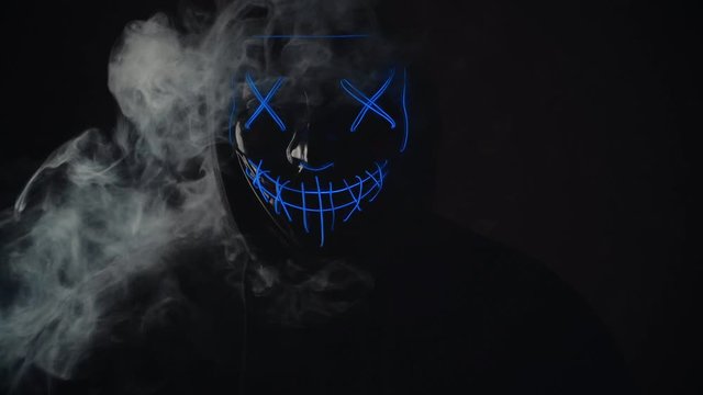 Man with lighting neon glow mask in hood and vape vapour from mask on black background. Halloween and horror concept