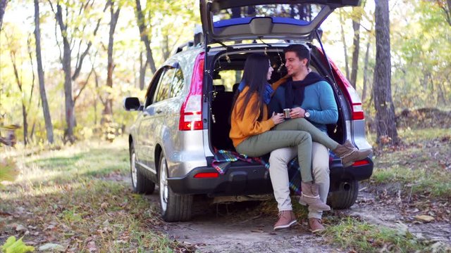 Young couple is sitting in car trunk with hot tea in metal cups and chatting and laughing. Autumn forest