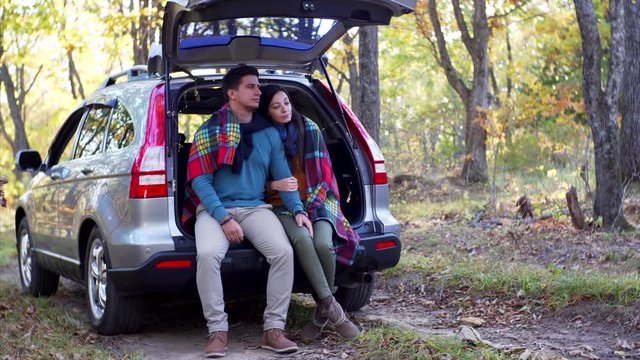 Young man and girl - romantic couple - are sitting in car trunk under plaid enjoying golden autumn in forest
