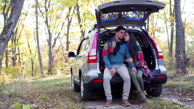 Young man and girl - romantic couple - are sitting in car trunk under plaid enjoying each other and golden autumn in forest