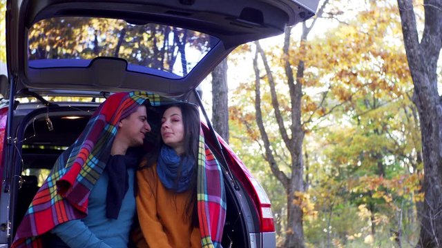 Young man and woman are enjoying each other in car trunk under warm plaid. Sunny autumn wood