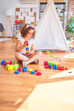 Beautiful toddler wearing glasses and unicorn diadem sitting playing with building blocks at kindergarten