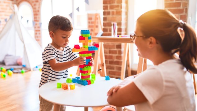 Beautiful teacher and toddler boy playing with construction blocks bulding tower at kindergarten