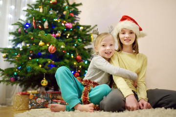 Two adorable sisters decorating a Christmas tree with colorful glass baubles at home. Family leisure at wonderful Xmas time.