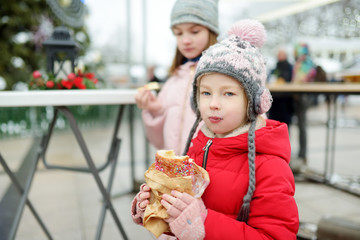 Two cute young sisters eating Czech trdelnik on traditional Christmas fair in Vilnius, Lithuania. Children enjoying sweets, candies and gingerbread on Xmas market.