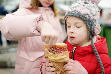 Two cute young sisters eating Czech trdelnik on traditional Christmas fair in Vilnius, Lithuania. Children enjoying sweets, candies and gingerbread on Xmas market.