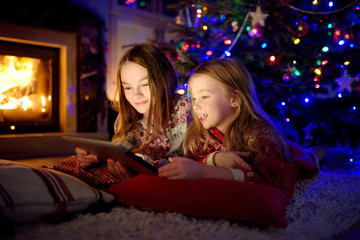 Two cute young sisters using a tablet pc at home by a fireplace in warm and cozy living room on Christmas eve.