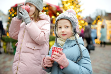 Two adorable sisters drinking hot chocolate on traditional Christmas fair in Riga, Latvia. Children enjoying sweets, candies and gingerbread on Xmas market.