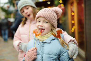 Fototapeta na wymiar Two adorable sisters having rooster-shaped lollipops on traditional Christmas fair in Riga, Latvia. Children enjoying sweets, candies and gingerbread on Xmas market.